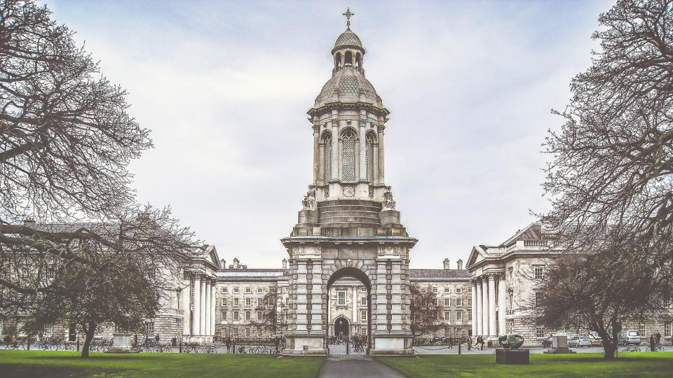 The front square of Trinity College Dublin during the daytime, on each edge of the photo are two large trees and between the trees stands a bell tower